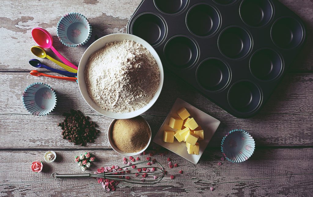 A selection of baking equipment including a cupcake tin, measuring spoons, bowls of sugar and flour, a whisk and some cubed butter.