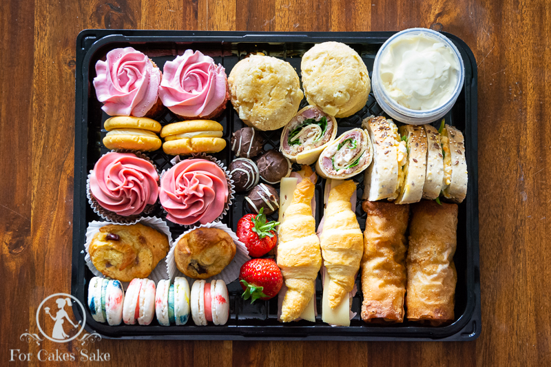 A For Cakes Sake Afternoon Tea Box. Box includes cupcakes, strawberry milkshake cake, custard shorties, cruffins, macarons, handmade chocolates, scones. It also has two sandwiches on seeded bread, two pin wheels, two mini croissants and two fajita chicken rolls. Garnished with two fresh strawberries.