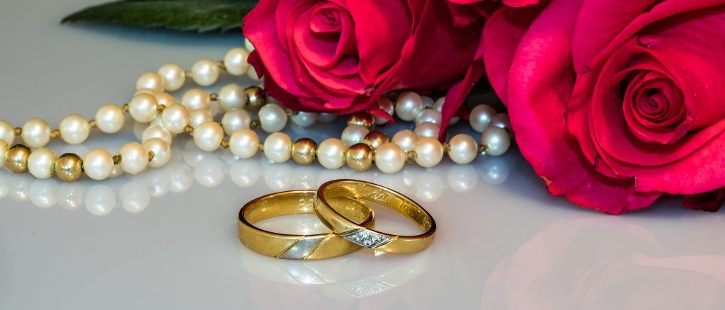 Two matching wedding rings beside red roses and a pearl necklace.